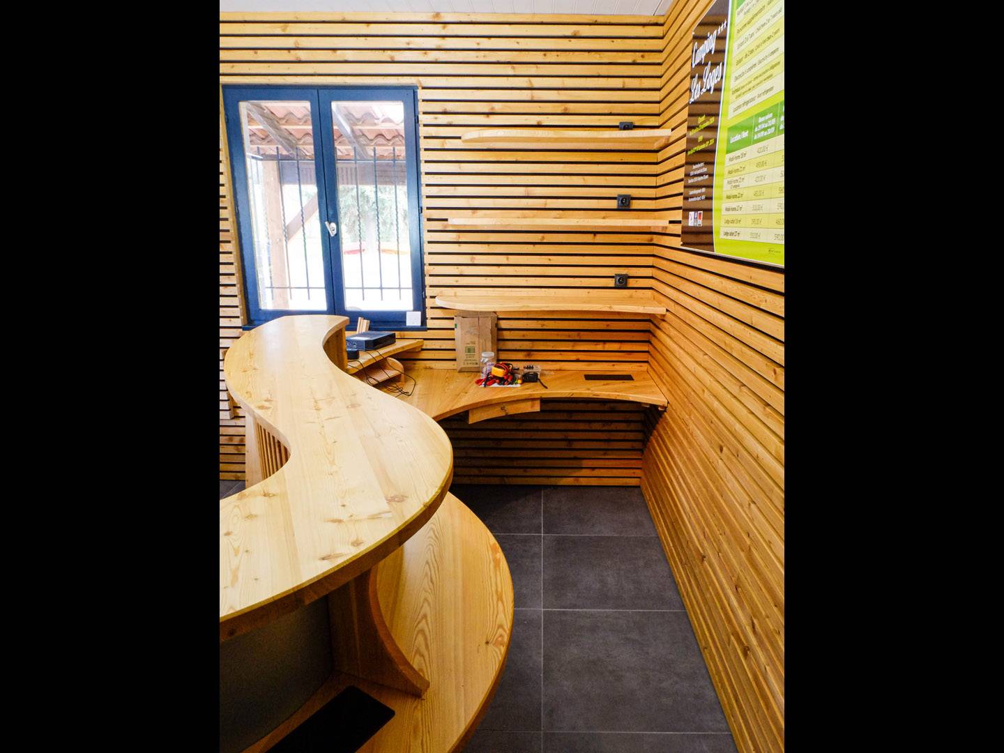 Projet global d'un accueil de camping. Comptoir d'accueil en courbe habillé en claire-voie avec des lattes de mélèze sur de la tôle laquée noire. Bureau pensé ergonomiquement. Tiroir caisse intégré. Atelier de la baleine. 2024